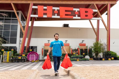 A person in a blue shirt carries red shopping bags outside an H-E-B store, surrounded by plants and garden supplies.