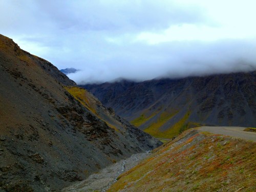 A scenic view of rugged mountains shrouded in clouds, with a winding road and colorful vegetation in the foreground.