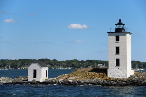 A white lighthouse stands on a rocky shore, with a small building nearby and boats in the background under a clear blue sky.