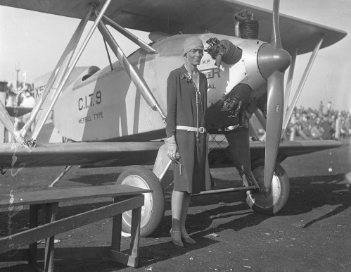 A woman in a vintage outfit stands beside a biplane, pointing confidently at the camera with a crowd in the background.
