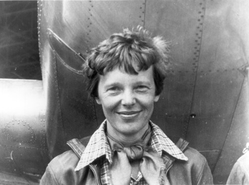 A smiling woman with short hair stands in front of an airplane, wearing a leather jacket and a scarf.