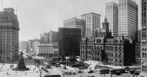 Historic black-and-white cityscape featuring buildings, streetcars, and pedestrians in a bustling urban square.