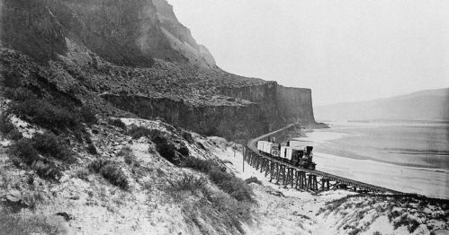 A vintage black-and-white photo of a train on a wooden trestle along a river, surrounded by rocky cliffs and vegetation.
