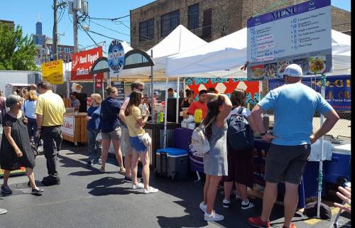 A busy outdoor market scene with people browsing food stalls under tents on a sunny day.
