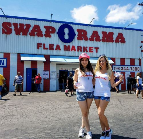 Two women in matching outfits pose in front of the Swap-O-Rama Flea Market, with a crowd and blue sky in the background.