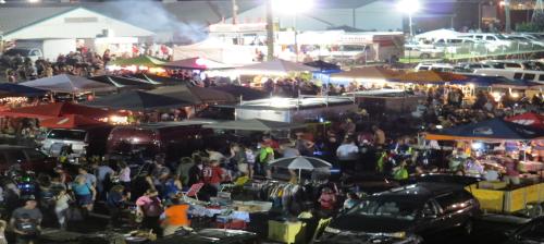 A bustling night market scene with crowds, food stalls, and colorful tents under bright lights.