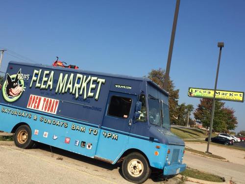 A blue food truck parked outside a flea market, with a sign indicating it's open on weekends from 8 AM to 4 PM.