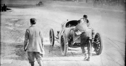 A vintage car being pushed by a man, with another man walking away on a dusty road.