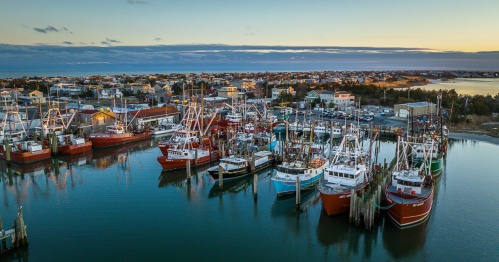 Aerial view of a harbor filled with fishing boats at sunset, with a coastal town in the background.