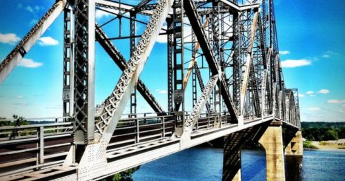 A metal truss bridge spans a river under a clear blue sky with scattered clouds.