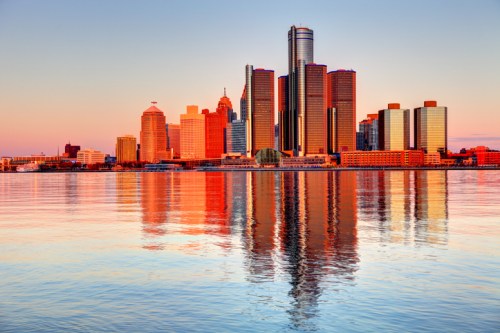 Sunset over a city skyline reflecting on calm water, with tall buildings and a colorful sky in the background.