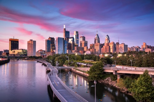 Philadelphia skyline at sunset, featuring modern buildings along the river with vibrant pink and purple clouds.