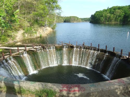 A circular concrete spillway with water cascading over the edge, surrounded by trees and a calm lake in the background.