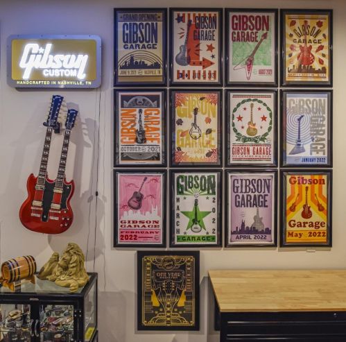 A wall display featuring framed posters of Gibson Garage events and two electric guitars in front.