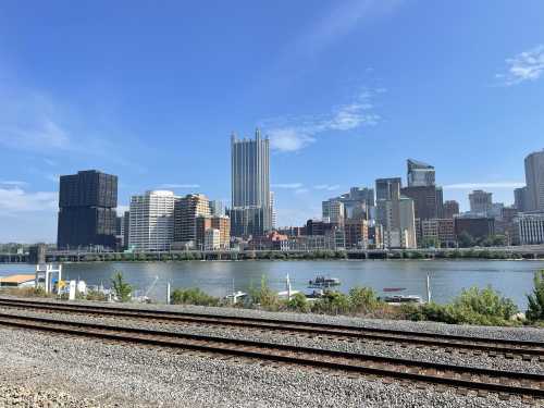 City skyline of Pittsburgh with tall buildings along the river, blue sky, and train tracks in the foreground.