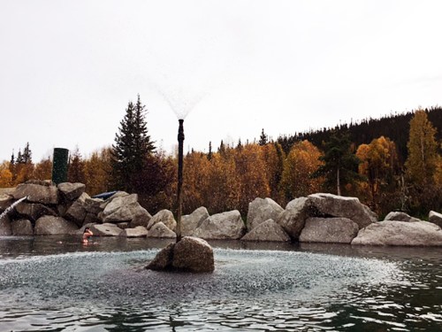 A serene hot spring with a fountain, surrounded by large rocks and autumn trees under a cloudy sky.