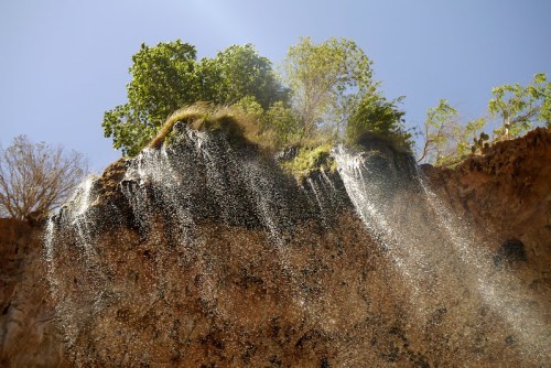 A waterfall cascading down a rocky cliff, with lush greenery at the top under a clear blue sky.