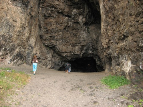 Two people approach a large cave entrance surrounded by rocky terrain and greenery.