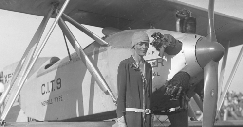A woman in vintage aviator attire stands beside a biplane, showcasing early aviation history.