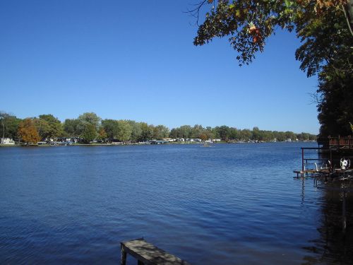 A calm lake scene with clear blue skies, trees lining the shore, and a wooden dock in the foreground.