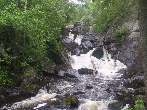 A cascading waterfall flows over rocky terrain, surrounded by lush green trees and foliage.