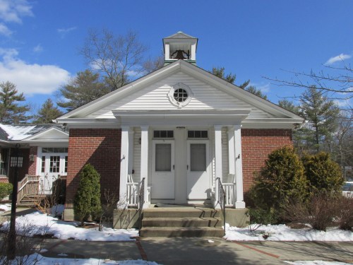 A white and brick building with a bell tower, surrounded by snow and trees under a blue sky.