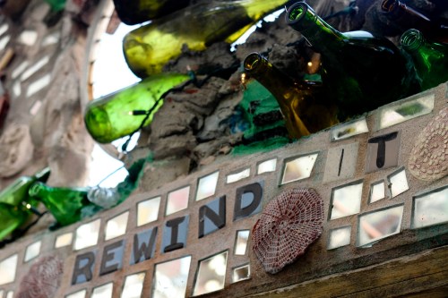 Colorful glass bottles and mosaic tiles form a decorative wall with the words "REWIND IT" prominently displayed.