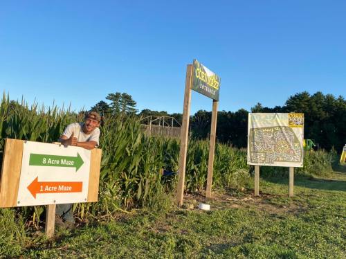 A person stands by signs for an 8-acre and 1-acre corn maze entrance, surrounded by tall corn plants.
