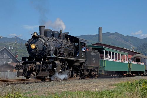 A vintage steam locomotive pulls a green passenger car along a scenic railway with mountains in the background.