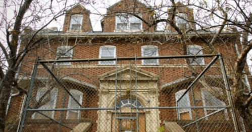 Abandoned brick building with a fenced entrance, surrounded by bare trees and a cloudy sky.