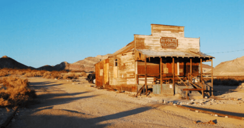Abandoned wooden building in a desert landscape, with mountains in the background and a clear blue sky.