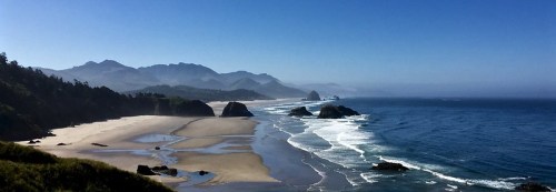 A scenic coastal view featuring sandy beaches, rocky formations, and distant mountains under a clear blue sky.