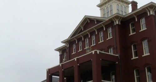 Historic red brick building with a tower, featuring multiple windows and a covered porch, under a cloudy sky.