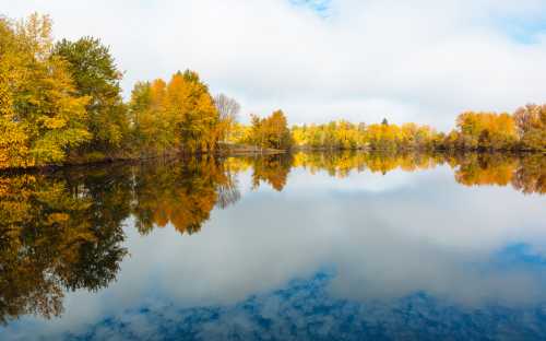 A serene lake surrounded by trees in vibrant autumn colors, reflecting the sky and foliage on the calm water.