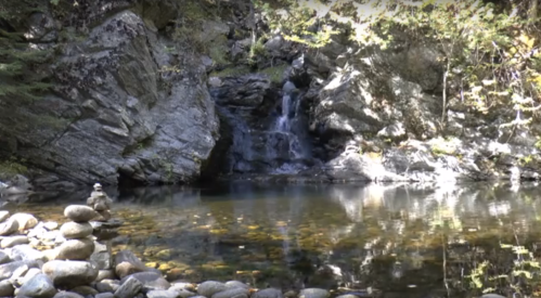 A serene scene featuring a small waterfall cascading into a calm pool, surrounded by rocks and autumn foliage.