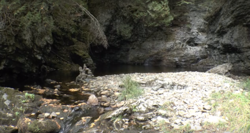 A serene rocky riverbank with a calm pool of water surrounded by lush greenery and steep stone walls.