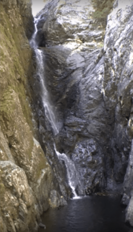 A cascading waterfall flows down rocky cliffs into a dark pool below, surrounded by rugged terrain.