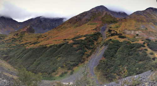 A panoramic view of rugged mountains with green slopes and patches of autumn foliage under a cloudy sky.