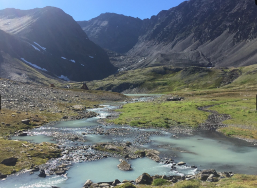 A serene mountain landscape with a winding stream, rocky terrain, and lush green patches under a clear blue sky.