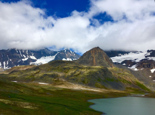 A scenic mountain landscape with rugged peaks, clouds, and a serene lake in the foreground.