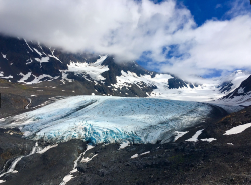 A vast glacier flows down a rocky mountain landscape, surrounded by snow-capped peaks and partly cloudy skies.