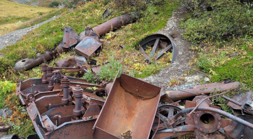 Rusty, abandoned machinery parts scattered on grassy terrain, surrounded by greenery and rocky ground.