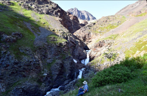 A person sits by a rocky riverbank, surrounded by lush green hills and mountains in the background.