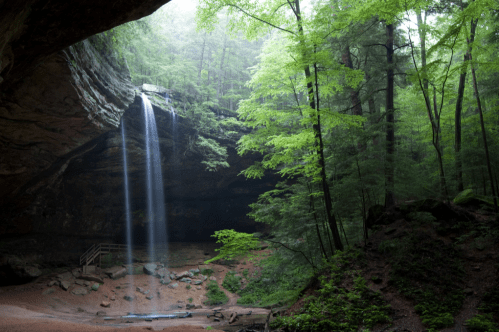 A serene cave scene with a waterfall cascading down rocks, surrounded by lush green trees and misty atmosphere.