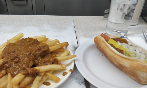A plate of fries topped with chili next to a hot dog with mustard and onions, accompanied by a glass of water.