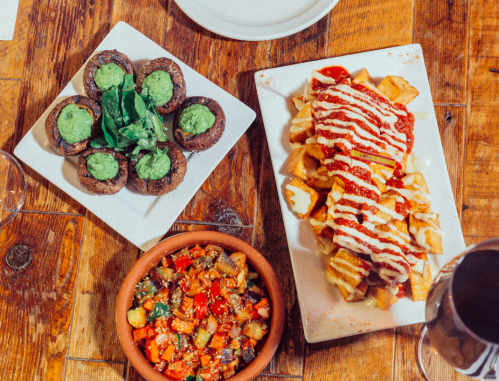 A wooden table with three dishes: stuffed mushrooms, loaded fries with sauce, and a colorful vegetable salad.