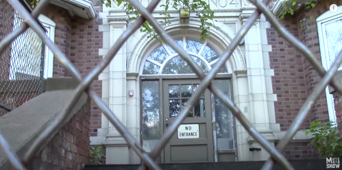 A brick building entrance behind a chain-link fence, featuring a "No Entrance" sign on the door.