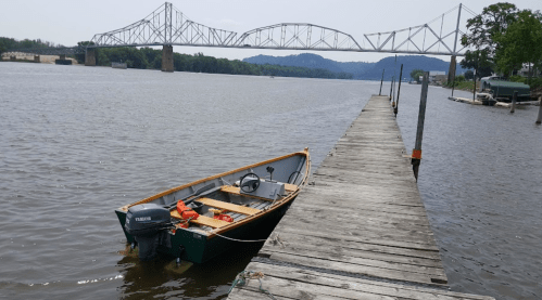 A small boat is docked at a wooden pier with a bridge spanning the river in the background.