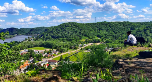 A person sits on a rocky ledge overlooking a lush green valley and a river, with clouds scattered in a blue sky.