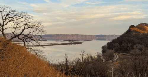 A serene landscape featuring a river, hills, and bare trees under a soft, cloudy sky.